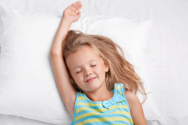 istock Good looking small female child with pleasant smile, stretches in bed, keeps eyes shut, dressed in casual nightclothes, closes eyes, sees pleasant dreams during afternoon rest, poses in white bed 1034229220