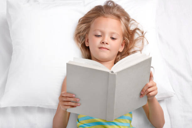 Pretty small preschooler reads attentively interesting story, holds opened book, lies in bed, enjoyes reading before sleep, has good rest. Clever little girl reads fairy tale. Hobby, children concept niños muy pequeños en edad preescolar lee con atención la interesante historia, sostiene el libro abierto, se encuentra en la cama, disfruta leyendo antes de dormir, tiene buen descanso. niña inteligente lee el cuento de hadas. hobby, concepto de los n
