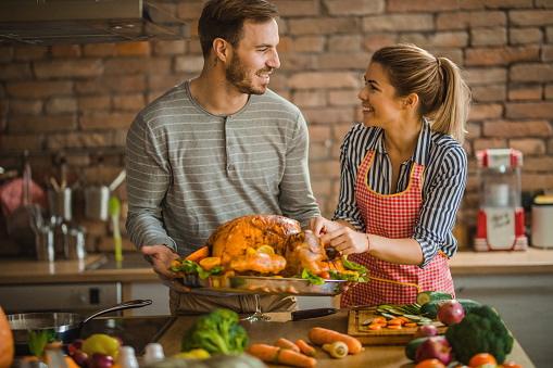 Young happy couple communicating while preparing Thanksgiving turkey for dinner at dining room.