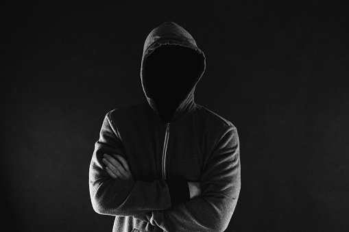 Anonymous and faceless man under hoodie with arms crossed isolated over dark background - incognito and mysterious criminal on internet activities concept