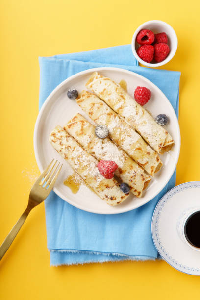 istock Rolled pancakes with blueberry and raspberry, caramel and powdered sugar on yellow background. Served with black coffee. Top view 1034235808