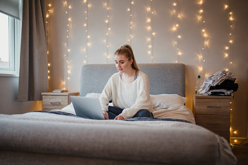 Young woman is sitting on her bed using a laptop.