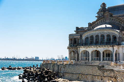 Color image depicting the old, derelict and abandoned casino on the Black Sea coast of Constanta, a port city in the south of Romania. People and tourists flock around the famous landmark, with the rough surf of the sea lapping against the banks.  The casino was built three separate times, with the first structure being erected of wood in 1880. It was designed to be a club and community center for elite and upper-class socialites willing to spend. Once considered Romania's Monte Carlo and a symbol of the City of Constanța, the most-recent and modern version was built in Art Nouveau style. Room for copy space.