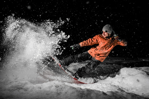 Young freeride snowboarder dressed in the orange sportswear and glasses jumping in powder snow at night