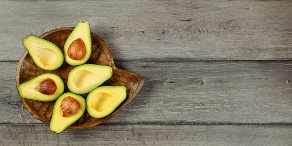 Tabletop view - avocado cut in half, arranged on wooden bowl, with dark grey wood desk under. Banner with space for text.