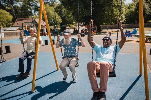 Three senior men are enjoying playing in a children's park in summer. They are on the swings together.