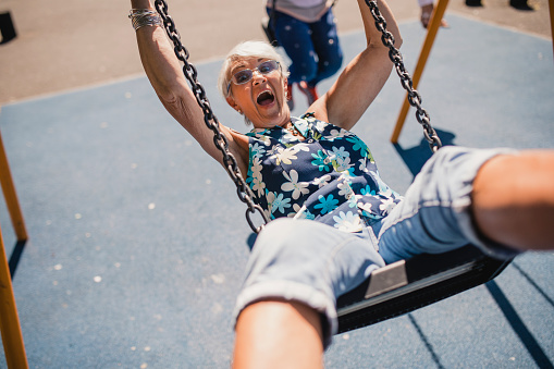 Senior woman on a child's swing in a public park. The sun is shining and she is laughing as she swings forward.