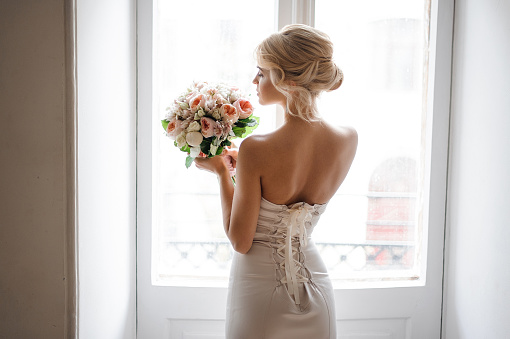 Back view of the elegant blonde bride dressed in a white dress holding a wedding bouquet