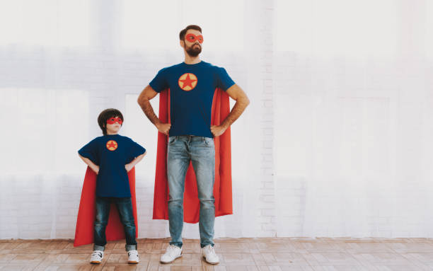 Father And Son In Superhero Suits. Family Concept. Father And Son. Red And Blue Superhero Suits. Masks And Raincoats. Posing In A Bright Room. Young Happy Family Holiday Concept. Resting Together. Save The World. Get Ready. Arms Akimbo. superhero photos stock pictures, royalty-free photos & images
