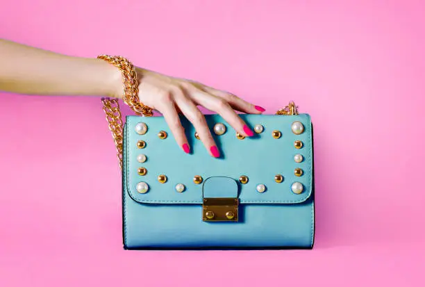 Photo of Sky blue handbag purse and beautiful woman hand with red manicure isolated on pink background.