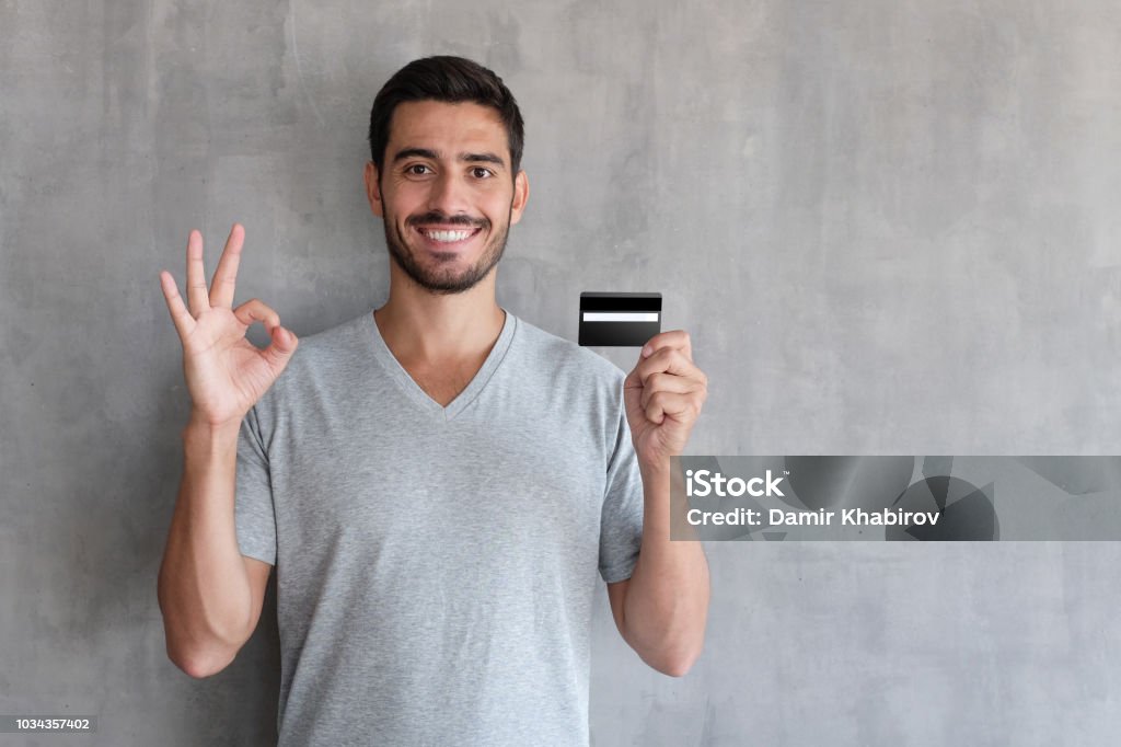 Closeup portrait du jeune beau caucasien guy illustration isolé sur fond gris, souriant, montrant OK signe avec sa main tout en maintenant la carte de crédit ou de débit dans un autre, sentiment satisfait - Photo de Carte de crédit libre de droits