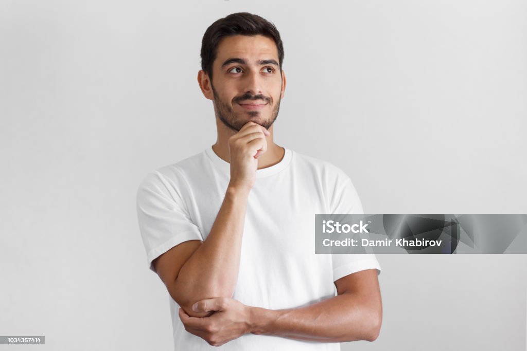 Horizontal shot of young European guy pictured isolated on grey background standing in blank T-shirt against wall, pressing fist to jaw as if dreaming of something, looking pensive and happy Contemplation Stock Photo