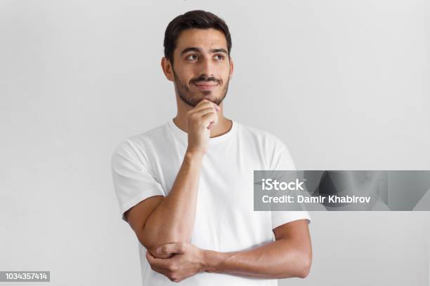Horizontal Shot Of Young European Guy Pictured Isolated On Grey Background Standing In Blank Tshirt Against Wall Pressing Fist To Jaw As If Dreaming Of Something Looking Pensive And Happy Stock Photo - Download Image Now