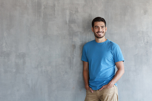 Indoor photo of handsome European guy pictured isolated against grey textured wall standing in blue T-shirt and trousers leaning to wall with shoulder, feeling confident and happy, enjoying free time