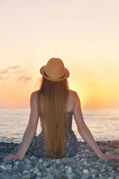 istock Girl in the hat sitting on the seashore. Sunset time. Back view 1034379372