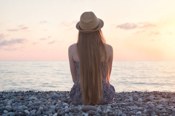 istock Girl in the hat sitting on the seashore. Sunset time. Back view 1034379410