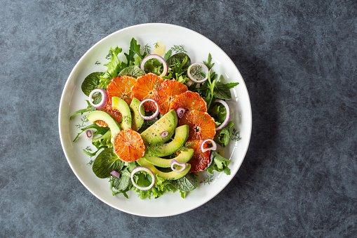 Avocado and blood orange salad with fennel, onions, and cracked pepper over mixed greens on a large white plate. This vegan salad is on top of a bluish gray table and shot top down.