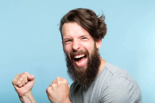 victory success and achievement. excited thrilled agitated guy making a winner gesture. hipster man portrait on blue background. emotional reaction and facial expression concept.