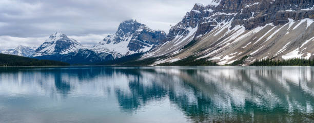 istock Bow Lake - A panoramic cloudy Spring day view of still snow-capped peaks, from left to right - Mt Hector, Bow Peak, BowCrow Peak, Crowfoot Glacier, and Crowfoot Mountain, reflecting in calm and colorful Bow Lake, Banff National Park, AB, Canada. 1034494900