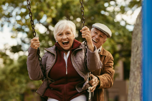 Senior man pushing his partner on swing Senior man pushing his female partner on swing in park and having fun together. Playful and happy senior couple enjoying at swing in park . swing stock pictures, royalty-free photos & images