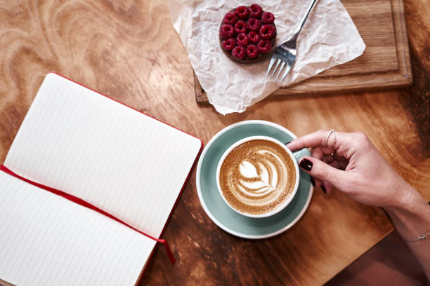 Cup of coffee latte on wooden table or background in woman hands from above. Having lunch in cafe. Opened notebook, space for design template.