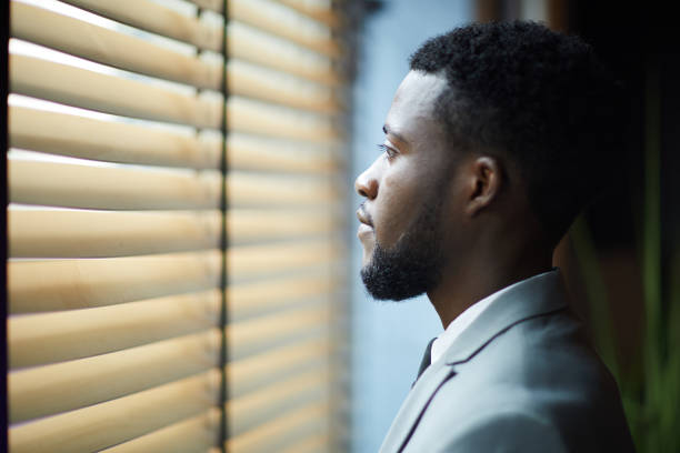 istock Pensive handsome young African man with beard looking through blinds while thinking about new project in corridor 1034581318