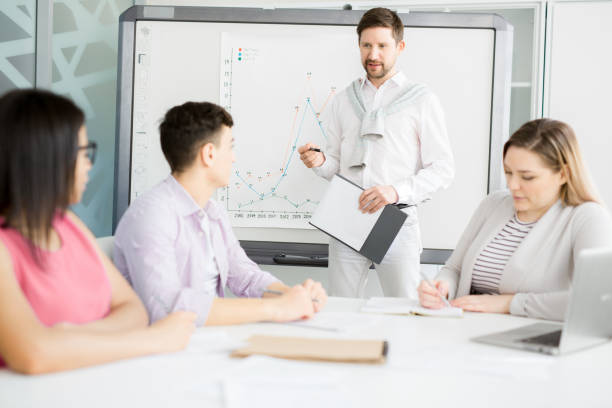 istock Content handsome young bearded manager in stylish outfit holding clipboard and looking at audience while explaining graph on whiteboard 1034584312