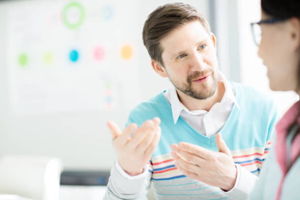 istock Content enterprising bearded manager wearing white shirt and sweater gesturing hands while explaining his idea to colleague in office 1034584426