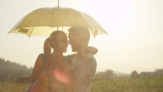 CLOSE UP, LENS FLARE: Happy Caucasian man and woman in love spin in the refreshing summer rain. Lovely blonde girl resting in smiling boyfriend's arms as they dance and spin around the rainy nature.