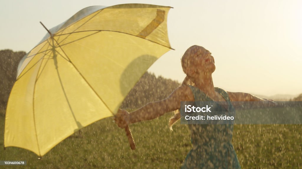 CLOSE UP: Carefree young blonde woman lets the spring rain fall down on her. CLOSE UP: Carefree young blonde woman lets the refreshing spring rain fall down on her. Cheerful girl in green sundress unwinds and puts her umbrella to the side and dances in the cool summer rain. Rain Stock Photo