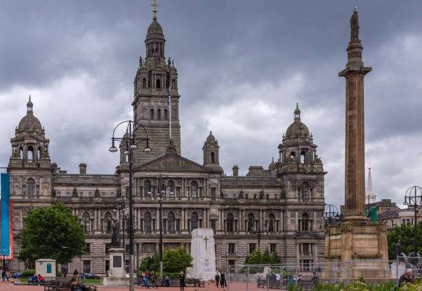 Glasgow, Scotland, UK - June 17, 2012: Brown stone Glasgow City Chambers building aginst dark cloudscape. Other monuments are Sir Walter Scott column, and war memorial. People of George Square.