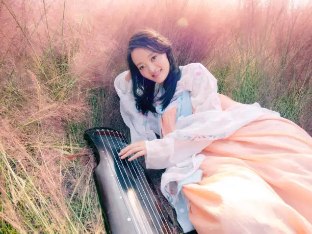 Portrait of beautiful young Chinese woman wearing ancient costume playing guqin in the pink hairawn muhly field.