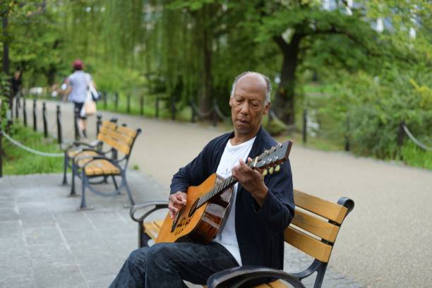A mature individual sitting at a park bench, playing the guitar on a late summer afternoon.
