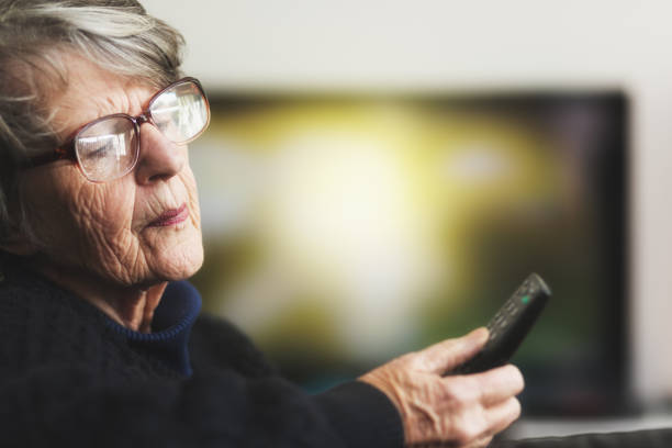 Senior woman holding TV remote can't read symbols A gray-haired woman in her 70s holds a TV's remote control, peering down at the markings on it but unable to read them clearly. read the fine print stock pictures, royalty-free photos & images