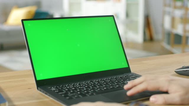 Man Uses Laptop with Green Mock-up Screen While Sitting at the Desk in His Cozy Living Room.