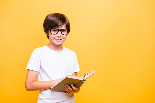Portrait of attractive young little cheerful school boy, smiling, wearing glasses reading a book over yellow background, isolated. Copy space