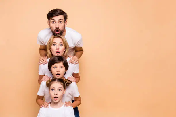 Photo of Adorable attractive surprised family, bearded father, blonde mother, boy and girl wearing white T-shirts, standing in odrer of hierarchy, holding hands on each other's shoulders. Copy space