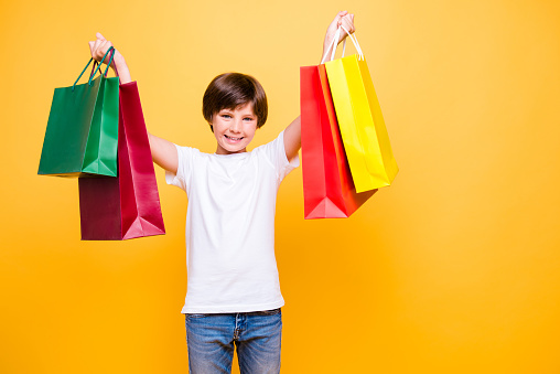 Portrait of attractive young cheerful school boy, smiling standing raising hands up with colorful bags over yellow background, isolated. Copy space