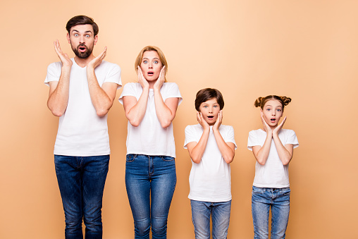 Portrait of young surprised family, bearded father, blonde mother and their little children wearing jeans and white T-shirts, standing straight and gesturing unsurely placing palms to cheeks