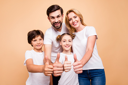 Portrait of young beautiful family, bearded father, blonde mother and their little children, boy and girl, wearing blue jeans and white T-shirts, showing thumbs up and smiling on beige background