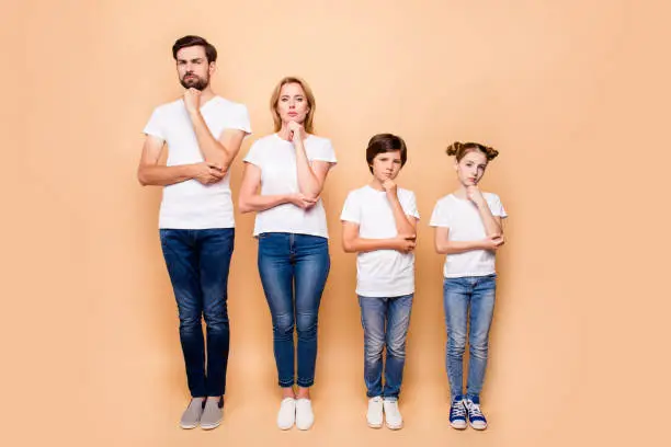 Photo of Full length portrait of beautiful  family bieng confused, bearded father, blonde mother and their children wearing jeans and T-shirts, standing straight showing uncertain gesture their hands to chins