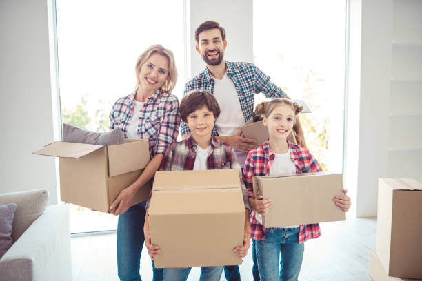 若い幸せな笑みを浮かべて家族 4 人カジュアルの立っているもののことを新しいアパートに移動光のリビング ルームで段ボール箱を持って身に着けています。 - people child twin smiling ストックフォトと画像
