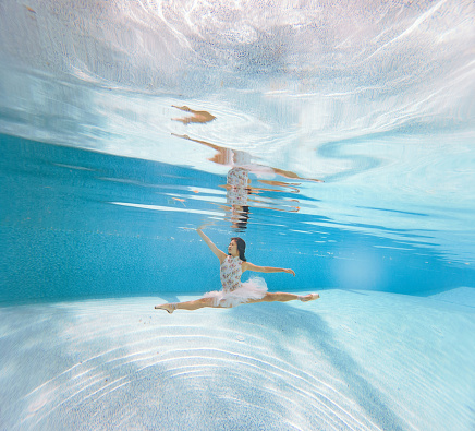 Japanese ballerina is dancing under water and making Grand Jete (twine). Her reflection is visible. Square image.