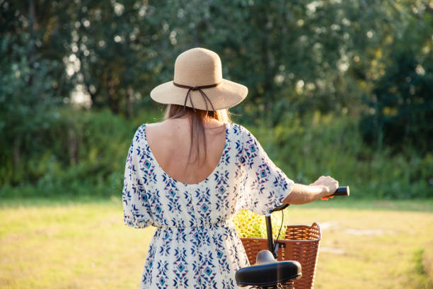 Rear view of an attractive woman walking along countryside with her bicycle. She is wearing light dress and a sun hat on a sunny summer afternoon.
