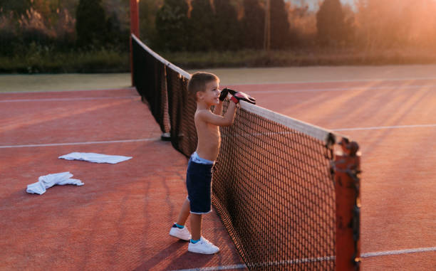 cute happy little boy standing in tennis playground leaned on tennis net . - tennis teenager little boys playing imagens e fotografias de stock