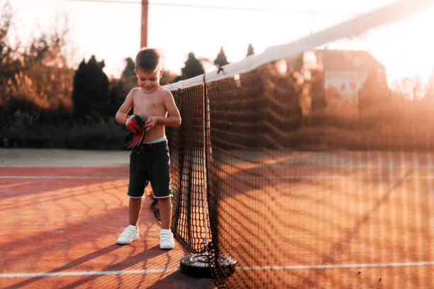 little boy standing on tennis playground and putting gloves on his hands. preparing him self for outside training. - tennis teenager little boys playing imagens e fotografias de stock