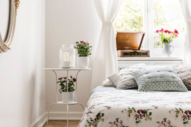 istock Rose loving woman's bedroom interior with a bed with flower pattern bedspread and pastel gray pillows. Red roses in pots on a stand by the bed. Real photo. 1034893474