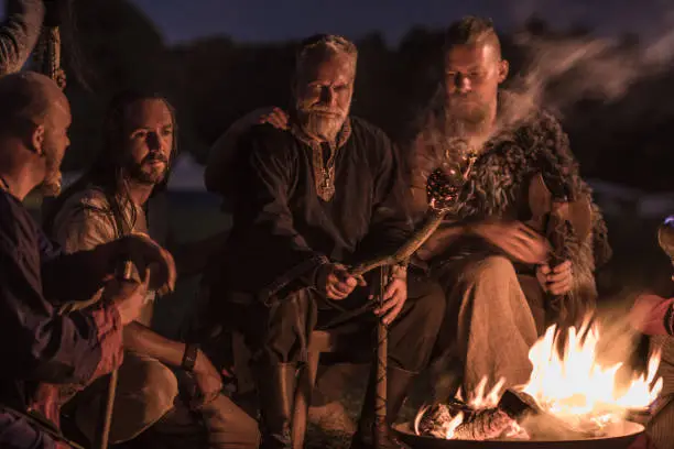 A hoard of Weapon wielding viking warriors sitting around a campfire in a battlefield scene in the countryside