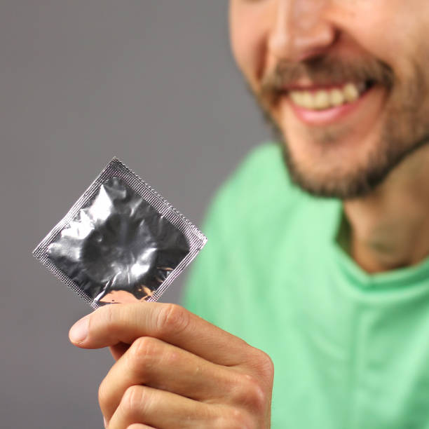 человек в зеленой рубашке держит презерватив в руке и улыбается, радость и уверенность в успехе, квадратная рамка - care condom confidence contraceptive стоковые фото и изображения