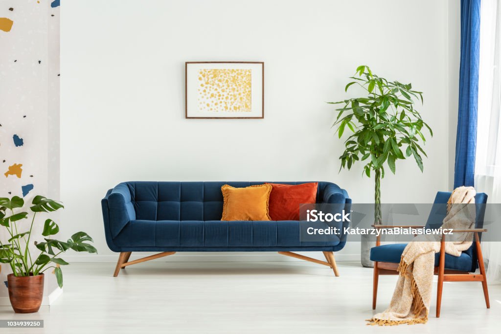 Mitte des Jahrhunderts moderner Stuhl mit einer Decke und einem großen Sofa mit bunten Kissen in ein geräumiges Wohnzimmer Interieur mit Grünpflanzen und weiße Wände. Echtes Foto. - Lizenzfrei Sofa Stock-Foto